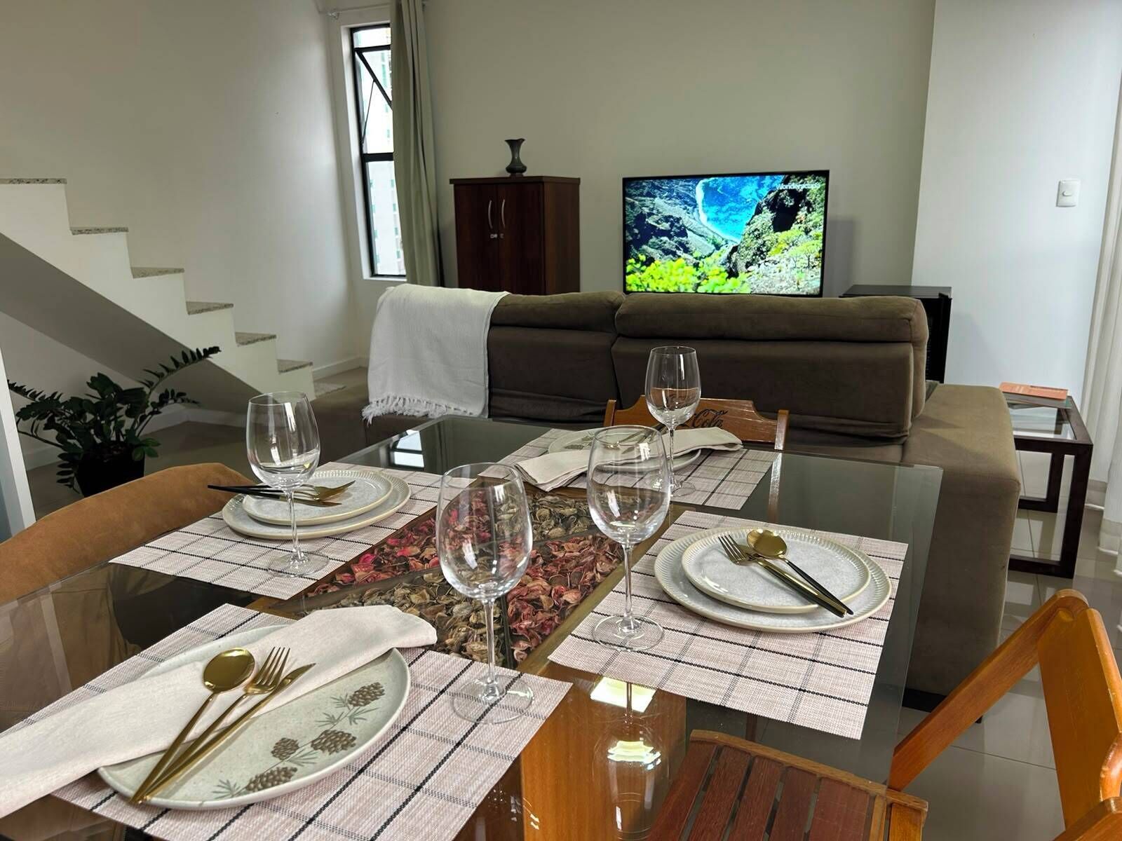 A dining area with a glass table set for four, a sofa, and a television in the background.