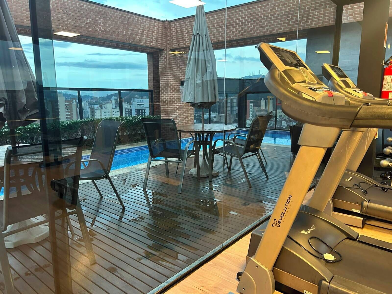A gym area with a treadmill, chairs, a table, and a view of a cityscape through large windows.