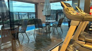 A gym area with a treadmill, chairs, a table, and a view of a cityscape through large windows.