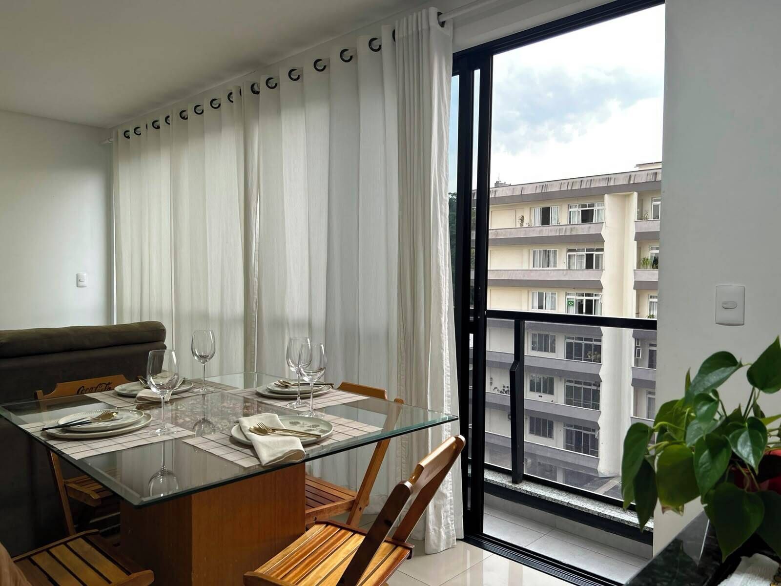 Comedor con mesa de vidrio, sillas y vista a un edificio residencial a través de un ventanal grande.