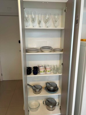 A kitchen cabinet with shelves containing wine glasses, plates, mugs, and cookware.