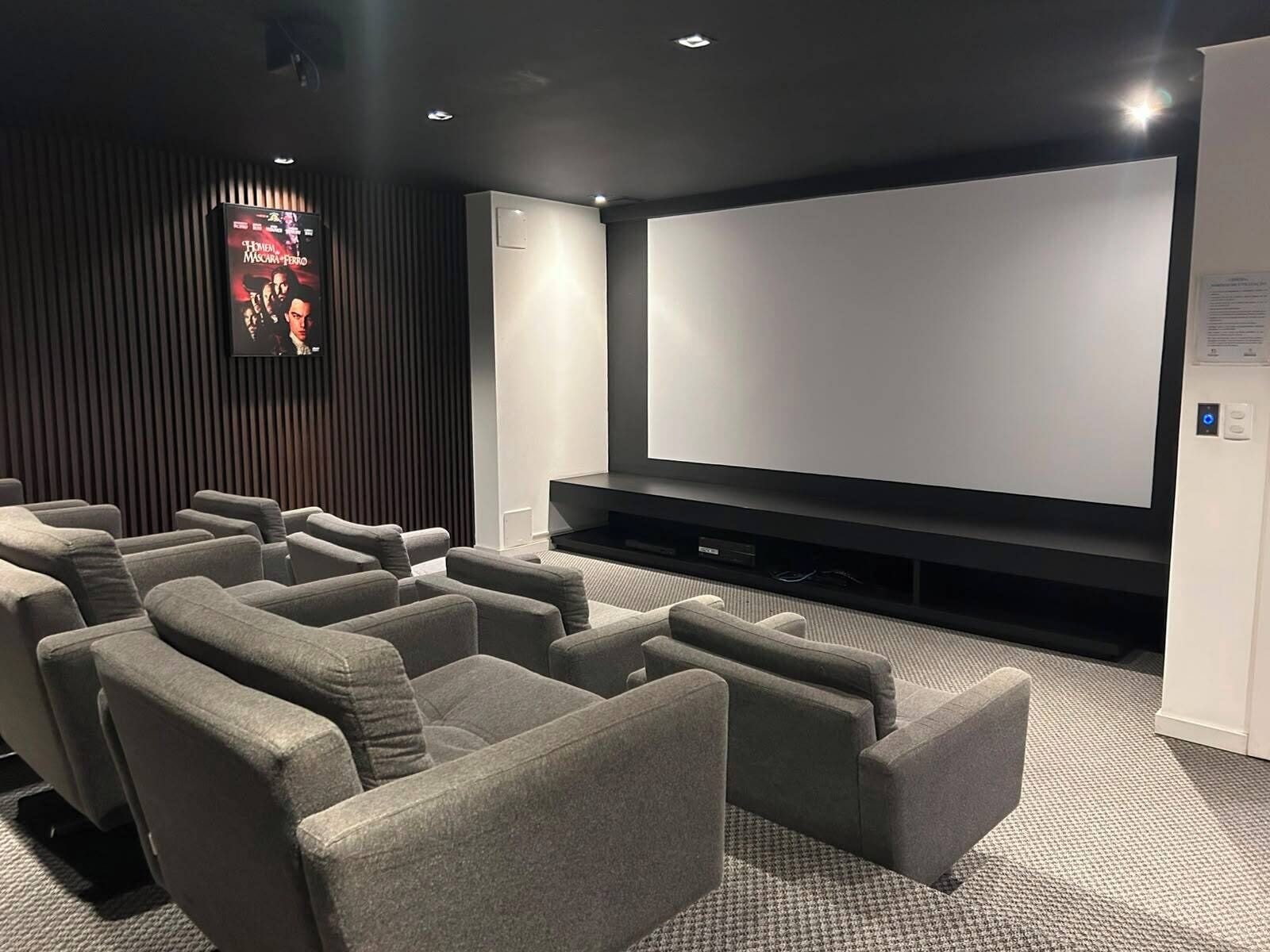 Uma sala de cinema com tela grande, assentos confortáveis e parede escura com linhas verticais.