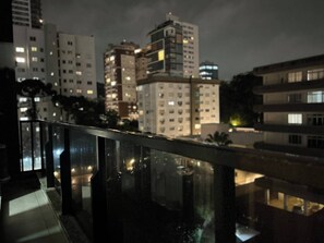 A nighttime view of a cityscape with high-rise buildings, a balcony railing, and a wet surface reflecting lights.