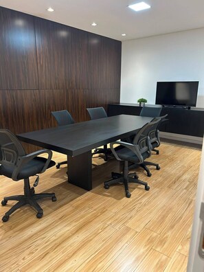 A conference room with a long table, four office chairs, a flat-screen TV, and a small plant on a cabinet.