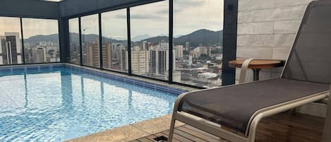 Uma piscina infinita no terraço com vista para a cidade, uma espreguiçadeira e um deck de madeira.