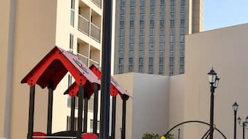 A playground with a slide and climbing structures, surrounded by a well-maintained lawn and a tall building in the background.