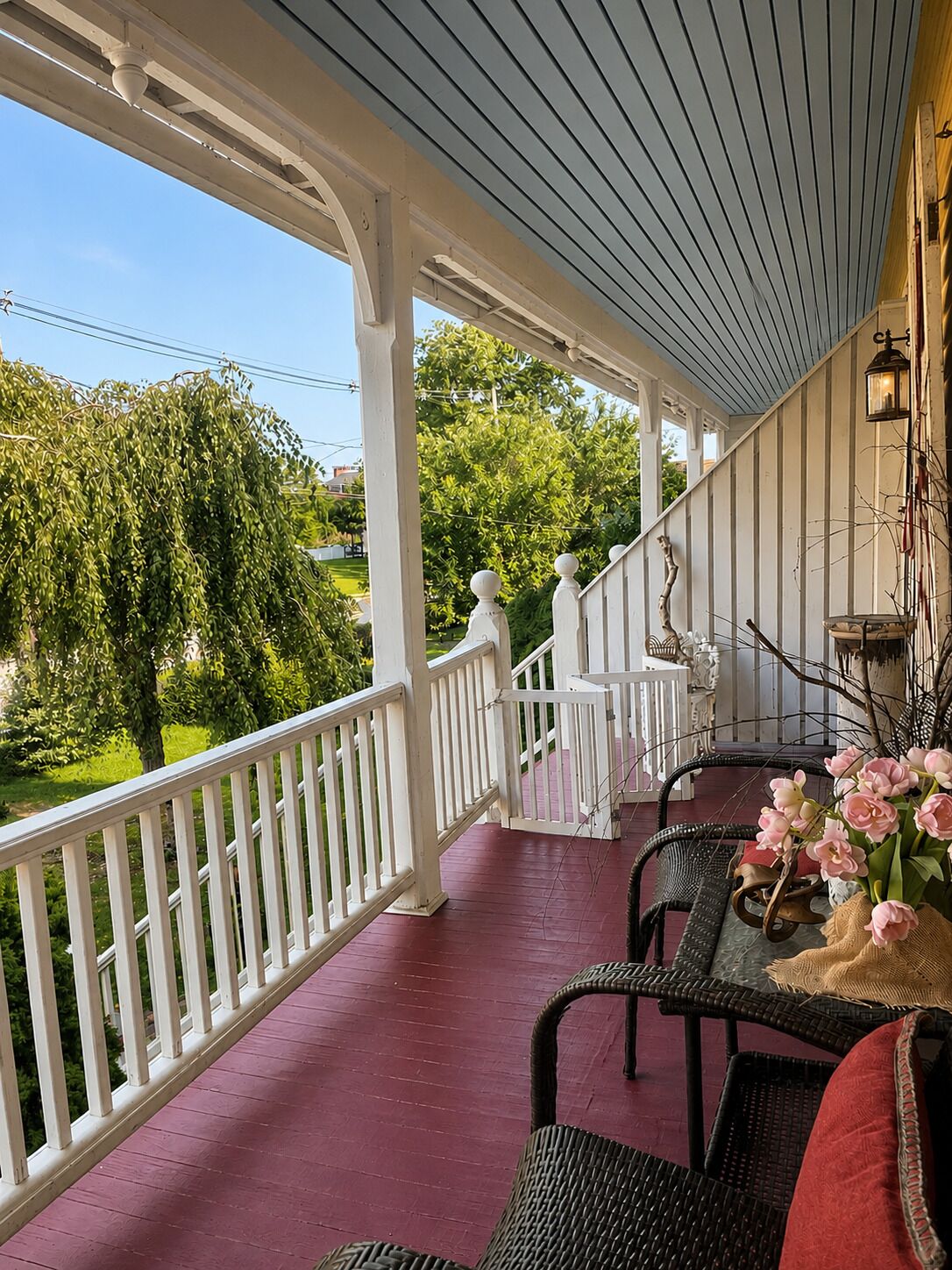 Eine überdachte Veranda mit Geländer, Korbmöbeln und einem Tisch mit Blumen.