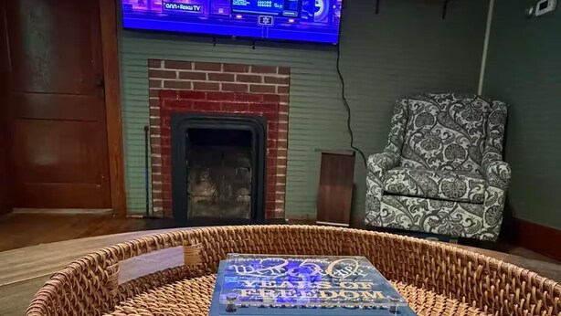 A living room with a television mounted on the wall, a fireplace, a patterned armchair, and a woven round seating area with American flag cushions.
