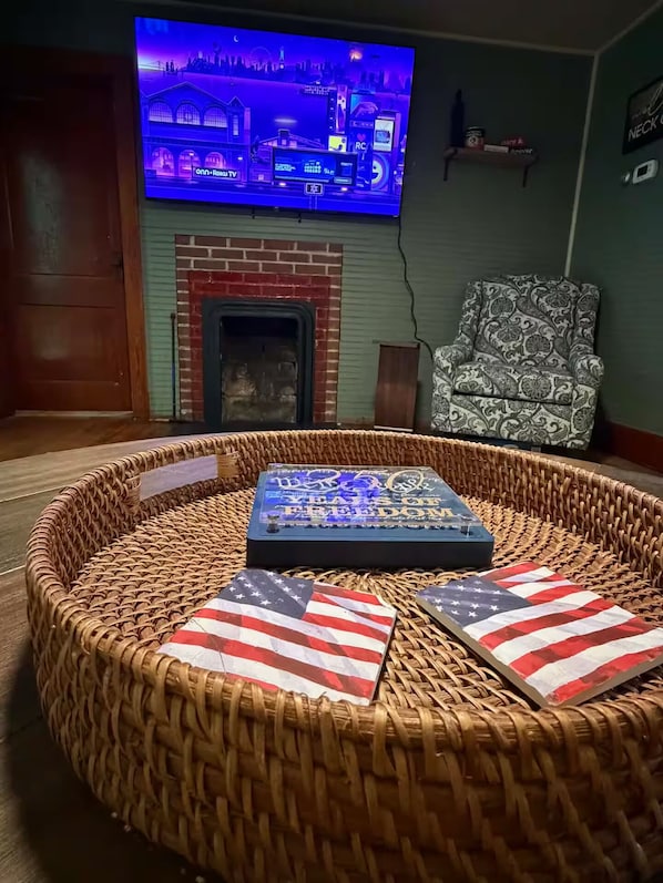 A living room with a television mounted on the wall, a fireplace, a patterned armchair, and a woven round seating area with American flag cushions.