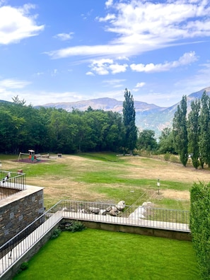 A grassy field with a playground and stone walls, surrounded by trees and mountains in the background.