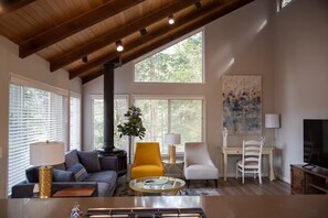A spacious living room with a high ceiling, large windows, a sofa, a yellow armchair, a white armchair, a glass coffee table, and a painting on the wall.