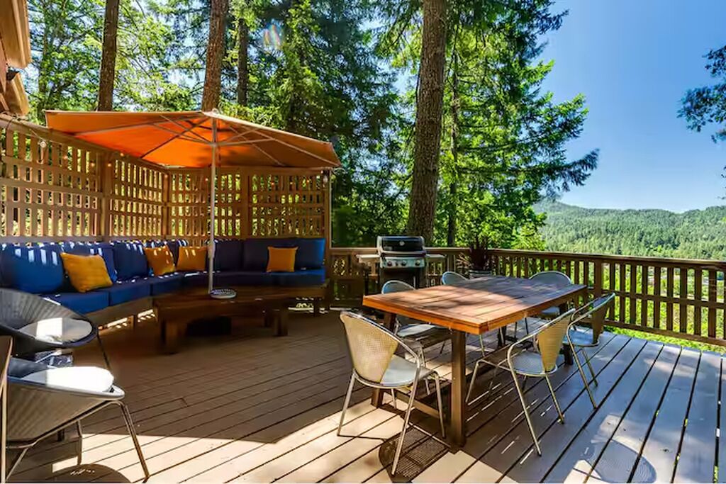Una terraza al aire libre con una mesa y sillas de madera, un sofá azul con cojines naranjas y una parrilla.