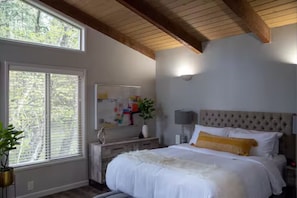 A bedroom with a large window, a bed with white bedding, a headboard, and a bedside table with a lamp.