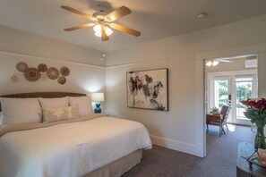 A bedroom with a bed, a ceiling fan, a wall-mounted artwork, and a door leading to a balcony with a chair and a potted plant. - C.H. Bailey House - Brenner Suite (Roseburg)