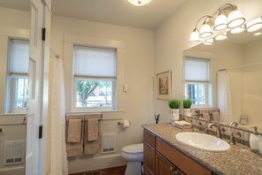 A bathroom with a white sink, vanity, and a large mirror. There are two windows with blinds, a towel rack, and a heater. - C.H. Bailey House - Brenner Suite (Roseburg)