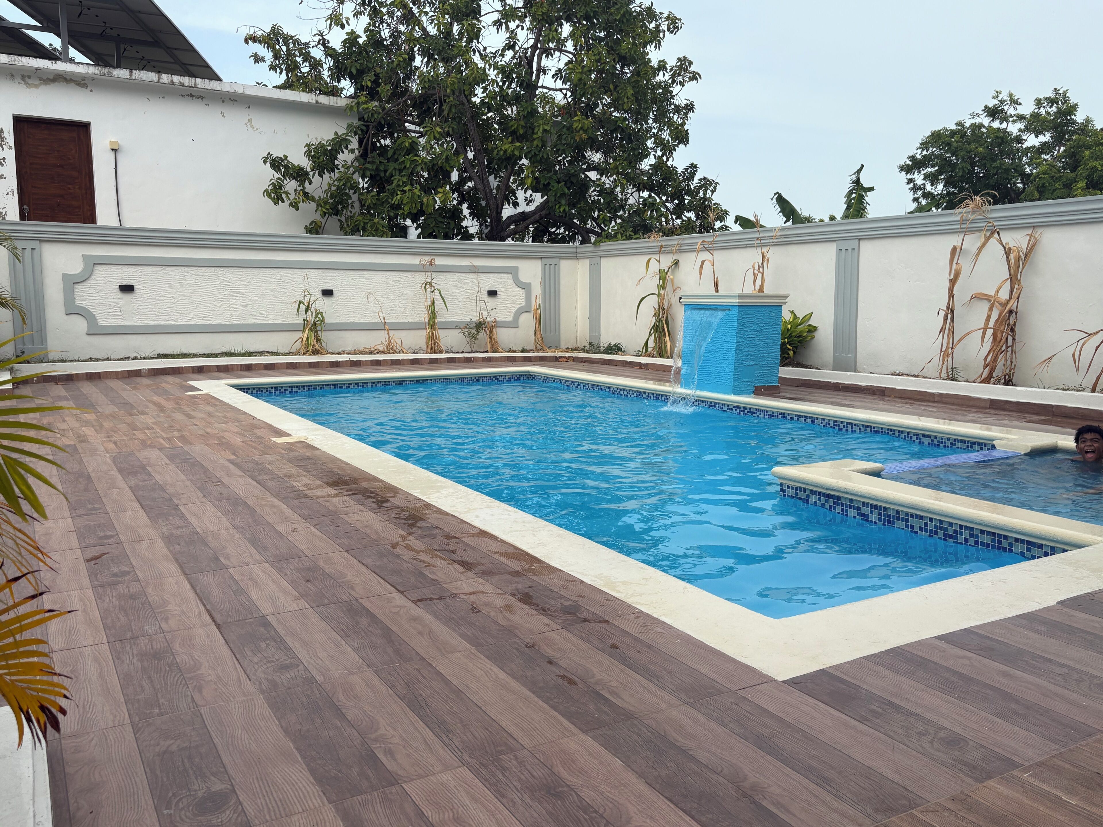 Una piscina rectangular con borde de azulejos, rodeada por una terraza de madera y una pared blanca con molduras decorativas.