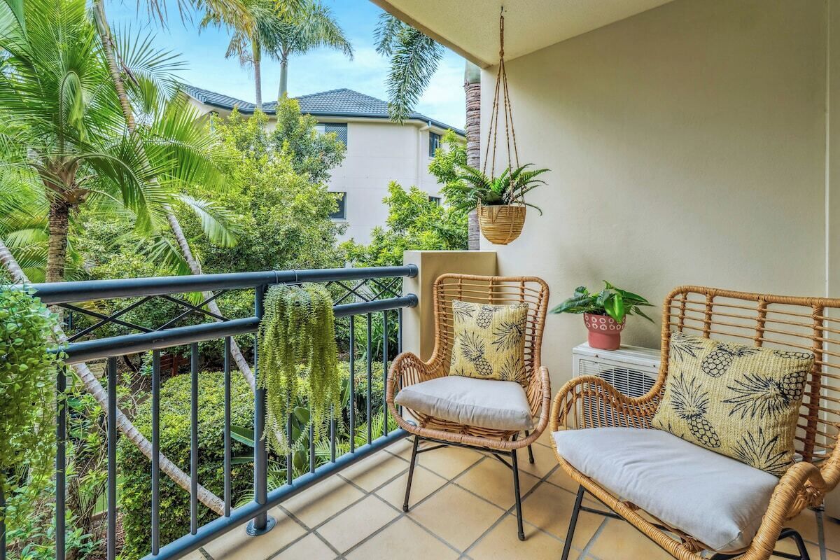 Un balcon avec des fauteuils en osier, une plante suspendue et une vue sur un immeuble et de la verdure.