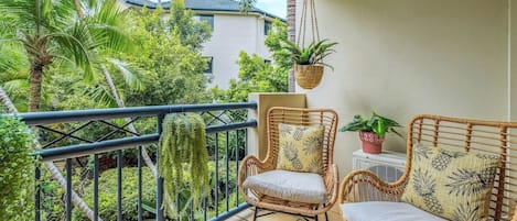 A balcony with wicker chairs, a hanging plant, and a view of a building and greenery.