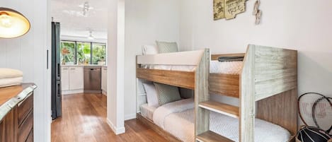 Een stapelbed met houten frame en wit beddengoed, een keuken met witte kastjes, en een eettafel met een lamp.
