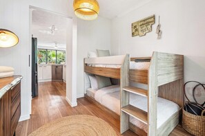 Un lit superposé avec une structure en bois et du linge de lit blanc, un coin cuisine avec des placards blancs et une table à manger avec une lampe.
