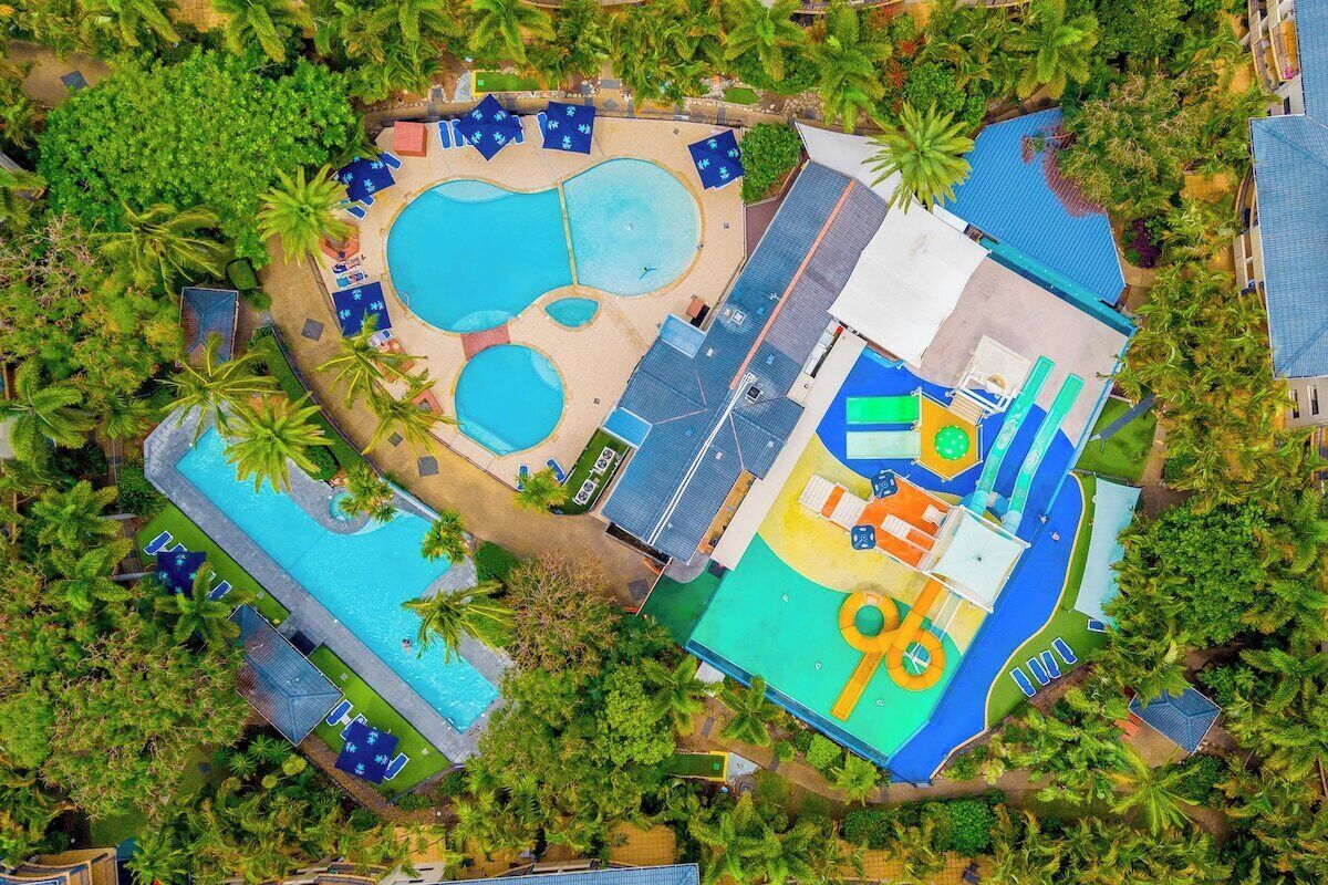 An aerial view of a resort with multiple swimming pools, a children's play area, and lush greenery.
