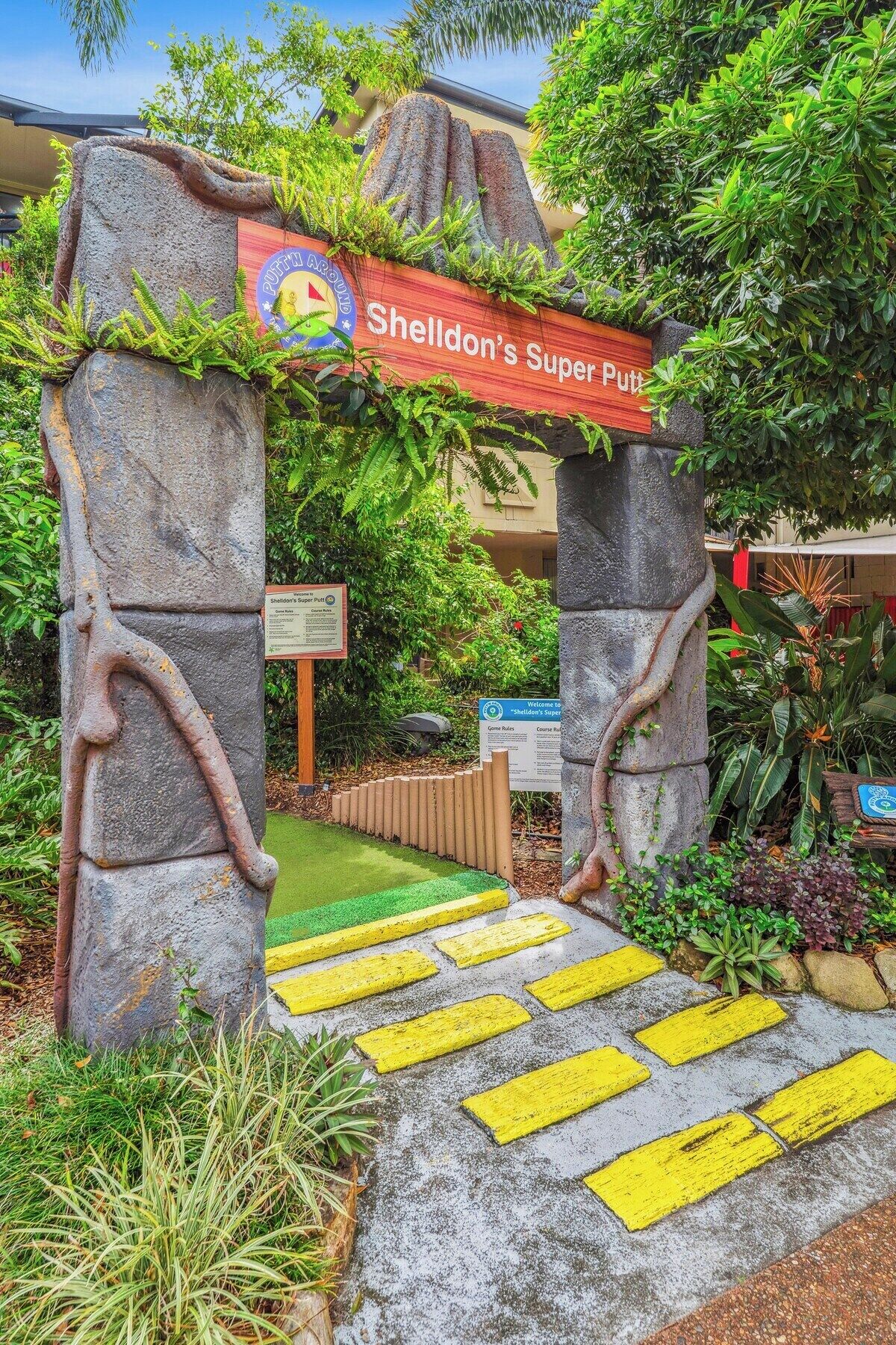 Ein steiner Torbogen mit einem Schild, auf dem 'Sheldon's Super Putt' steht, der zu einem Weg führt.