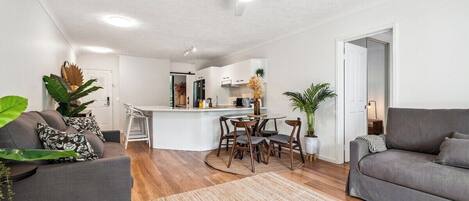 Uma sala de estar moderna com um sofá cinzento, uma área de jantar com mesa redonda e uma cozinha ao fundo.