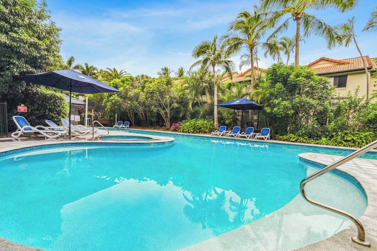 A swimming pool with lounge chairs and umbrellas, surrounded by palm trees and a building with a tiled roof.
