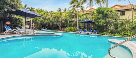 A swimming pool with lounge chairs and umbrellas, surrounded by palm trees and a building with a tiled roof.