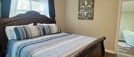 Un dormitorio con cama, ventilador de techo, ventana con cortinas y baño visible a través de una puerta abierta.