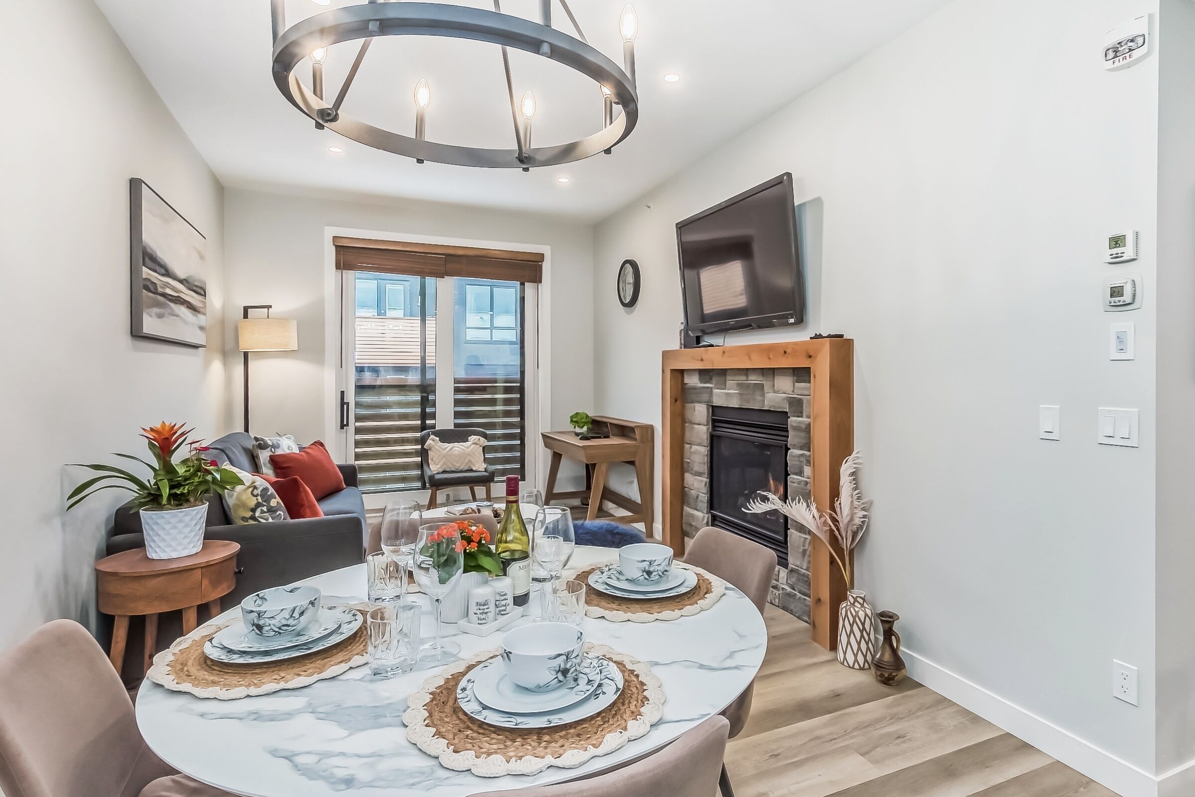 A dining area with a round table set for four, a television above the fireplace, and a large window with blinds.