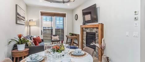 A dining area with a round table set for four, a television above the fireplace, and a large window with blinds.