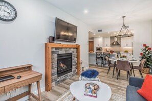 A modern living room with a fireplace, a television mounted above it, and a dining area with a chandelier. - Spring Creek Getaway, Luxury 1BR Condo in Canmore (Canmore)