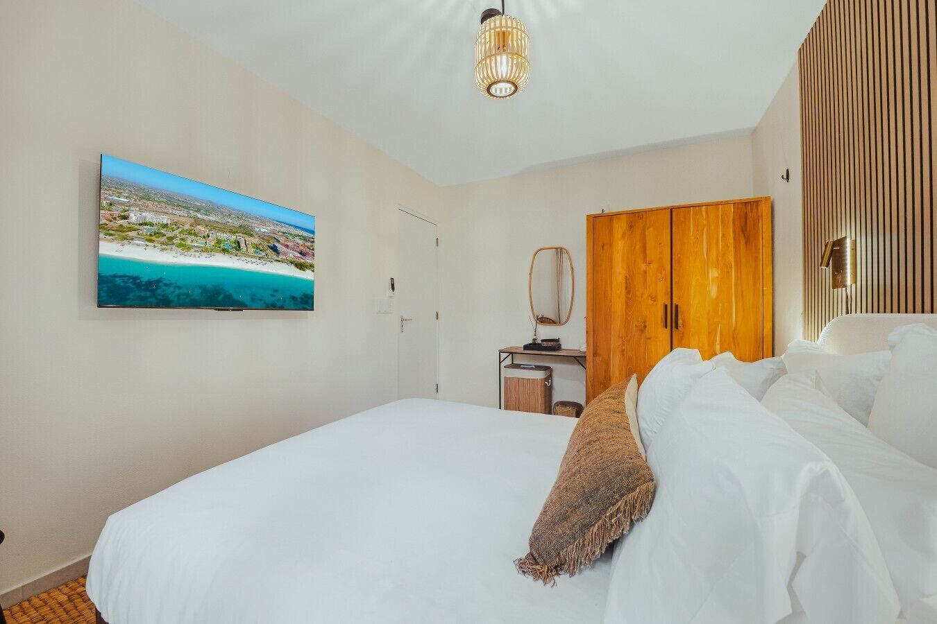 Quarto de hotel com cama, guarda-roupa de madeira, escrivaninha com espelho e televisão fixada na parede exibindo uma cena de praia.
