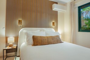 A hotel room with a bed, a nightstand, a lamp, an air conditioning unit, and a window with a view of greenery. - Coco Cottage Tropical 1BR Near Palm & Eagle Beach (Noord)