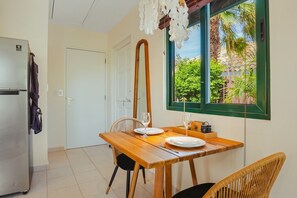 A compact kitchen with a refrigerator, a dining table with chairs, and a window with a view of greenery. - Coco Cottage Tropical 1BR Near Palm & Eagle Beach (Noord)