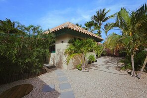 Une maison avec un toit en tuiles, entourée de palmiers et un chemin de gravier menant à l’entrée.