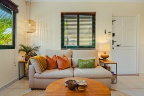 A living room with a sofa, a coffee table, a lamp, and a window with blinds. - Coco Cottage Tropical 1BR Near Palm & Eagle Beach (Noord)
