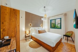 A modern bedroom with a wooden bed frame, a bedside table with a lamp, a window with a view of greenery, and a wall-mounted TV. - Coco Cottage Tropical 1BR Near Palm & Eagle Beach (Noord)