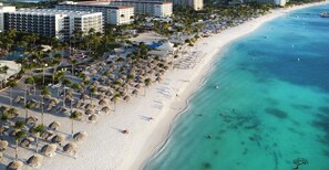 A beach resort with multiple hotel buildings, a sandy beach lined with palm trees and thatched umbrellas, and clear turquoise waters. - Coco Cottage Tropical 1BR Near Palm & Eagle Beach (Noord)