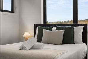 A neatly made bed with a white comforter and pillows, a bedside lamp, and a window with a view of the landscape. - Premium 3BR Snowy Mountains Retreat w/ Ducted A/C! (Jindabyne)