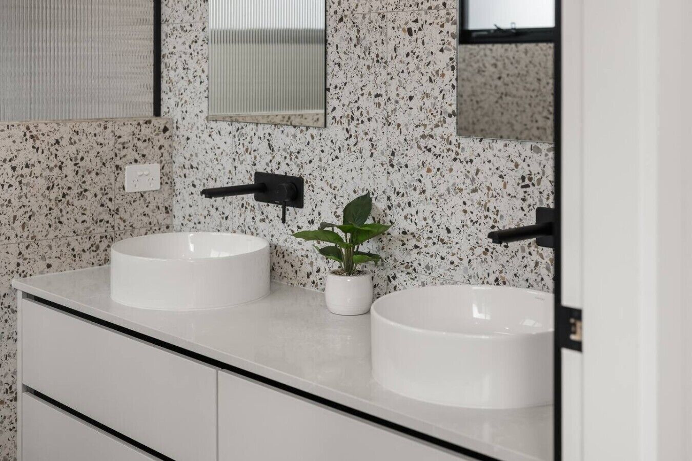 A bathroom with two white sinks, a small potted plant, and a mirror with a black frame.