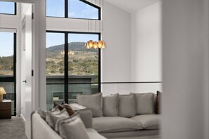 A modern living room with a sofa, a hanging light fixture, and a view of a hilly landscape through large windows. - Snowy Mountains Retreat with Indoor Fireplace (Jindabyne)