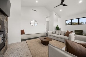 A modern living room with a stone fireplace, a round mirror, a sofa set, and a ceiling fan. - Snowy Mountains Retreat with Indoor Fireplace (Jindabyne)