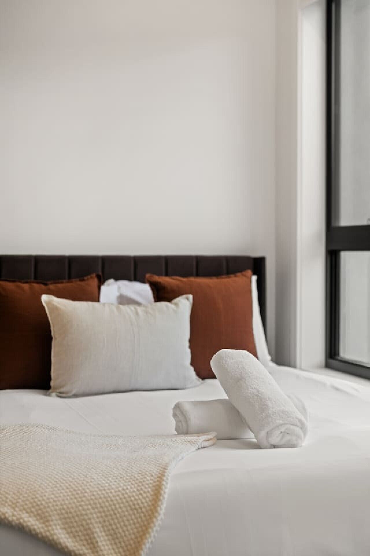 A neatly made bed with white linens, a brown headboard, and a window with a view.