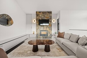 Ein modernes Wohnzimmer mit Kamin aus Stein, einem flachbildfernseher an der Wand, einem runden Couchtisch und einer Couch mit Kissen.