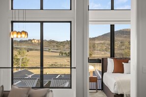 A room with large windows, a bed with pillows, a bedside table, and a hanging light fixture. - Stylish 3BR Snowy Mountains Getaway w/ Ducted A/C! (Jindabyne)