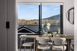 A dining area with a table and chairs, a window with a view of a hilly landscape, and a potted plant on the windowsill. - Stylish 3BR Snowy Mountains Getaway w/ Ducted A/C! (Jindabyne)