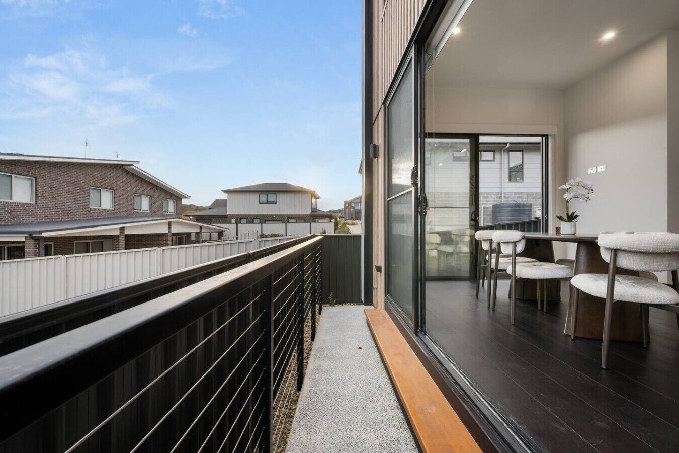 Een modern balkon met uitzicht op omliggende gebouwen, een glazen deur naar een eetgedeelte met stoelen en een tafel, en een kleine potplant.