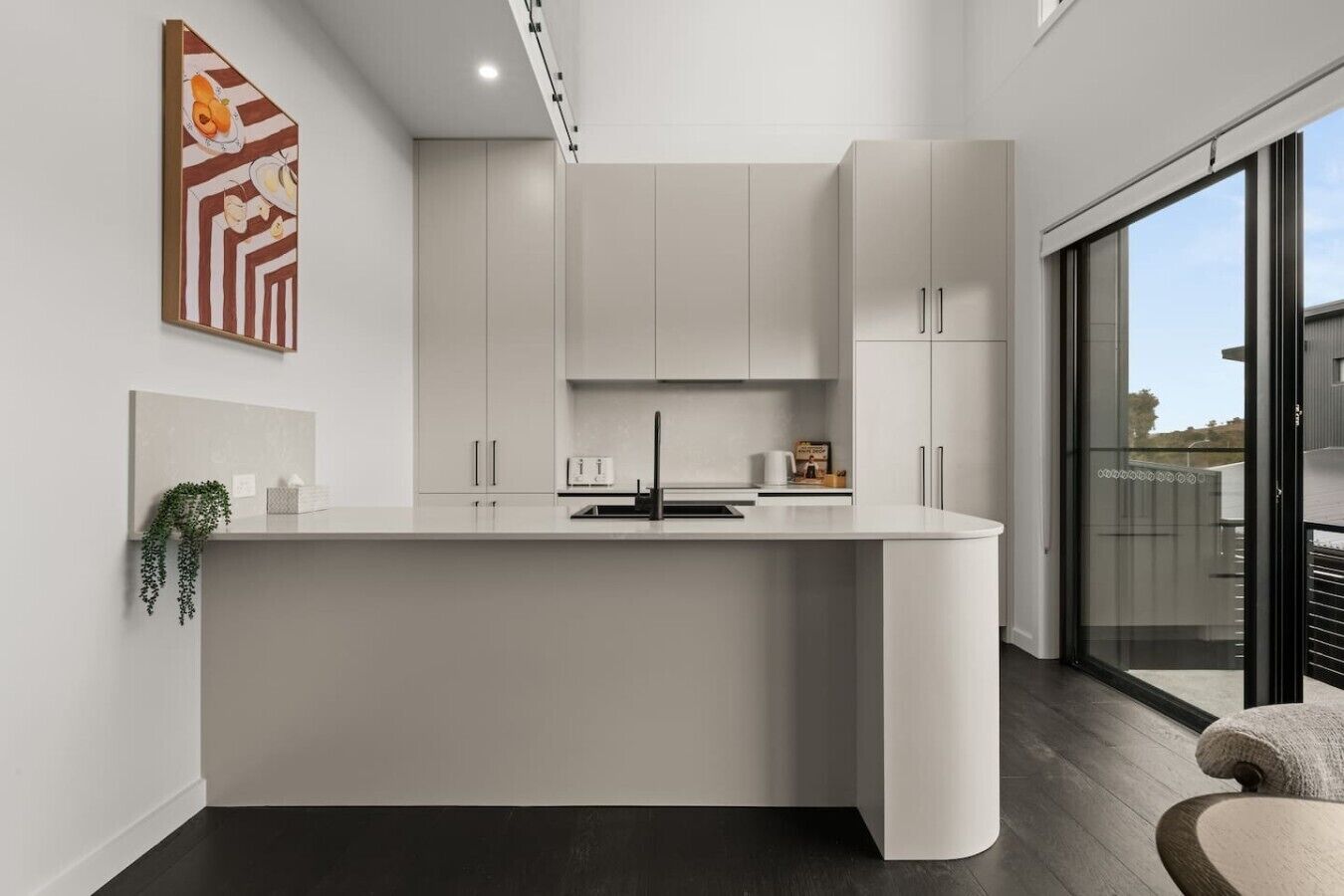 Une cuisine moderne dotée d’un îlot central aux lignes courbes, de meubles blancs et d’une grande fenêtre offrant une vue dégagée.
